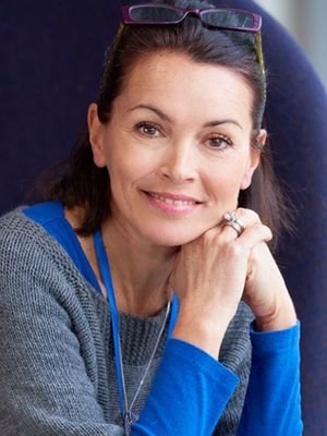 Paloma Silva - Vidente de Confianza Retrato de la vidente Paloma Silva sonriendo y apoyando la barbilla en sus manos, con un suéter gris y una camiseta azul, transmitiendo confianza y cercanía.